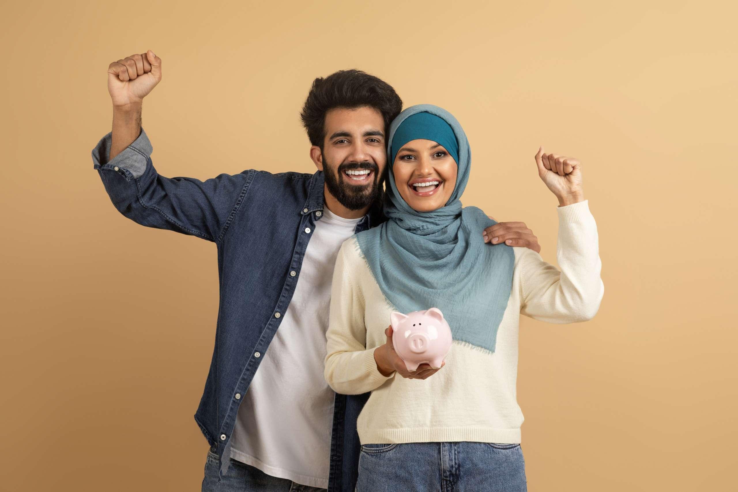 Happy Young Muslim Couple Holding Piggy Bank And Celebrating Success